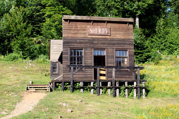 Old wooden sheriffs office with porch and sign stands elevated on posts in a grassy clearing,...