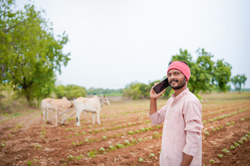 young indian farmer standing at agricultural field talking on smartphone