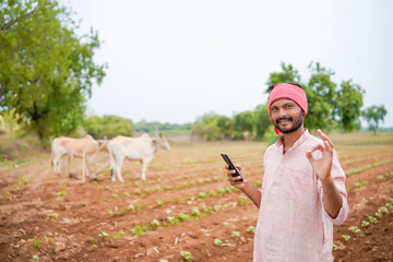 young indian farmer holding smartphone standing in agriculture field