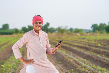 young indian farmer holding smartphone standing in agriculture field
