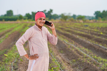 young indian farmer standing at agricultural field talking on smartphone