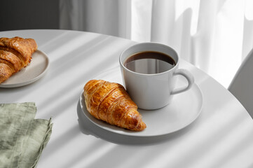 Cup of espresso coffee and fresh croissant on a plate on white table near window with morning shadows.