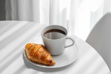 Cup of espresso coffee and fresh croissant on a plate on white table near window