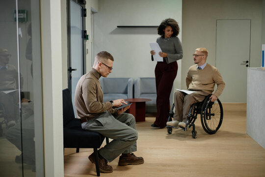 Caucasian young adult man sitting on bench using tablet, Black woman standing holding documents, colleague with disability in wheelchair talking