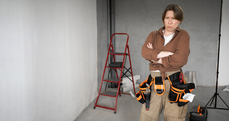 Unhappy repairwoman with tool belt in living room interior doing a home renovation, crossed arms...