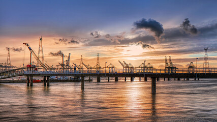 Long pier in the Scheldt with cranes in the background during a colorful sunset and dramatic cloud formations