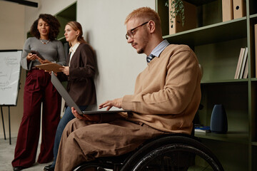 Caucasian young adult man with disability using laptop in wheelchair, two women standing in...