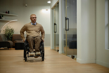 Caucasian man with disability using wheelchair moving through modern office corridor, wearing glasses and neutral expression, indoor business environment