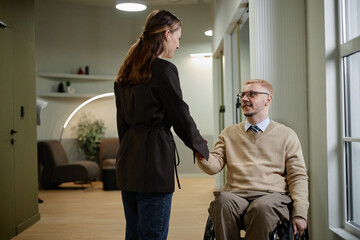 Caucasian man with disability sitting in wheelchair shaking hands with young adult woman standing...
