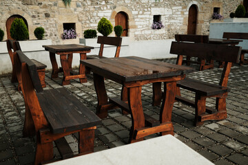 Rustic wooden benches and tables arranged in a stone-paved courtyard against a medieval stone wall with arched doors and flowers. Serbia country.