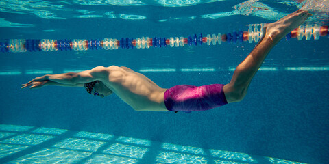 A male swimmer gracefully glides underwater in a swimming pool as sunlight casts intricate patterns on the pool floor. Concept of aquatic sports and natural light reflections.
