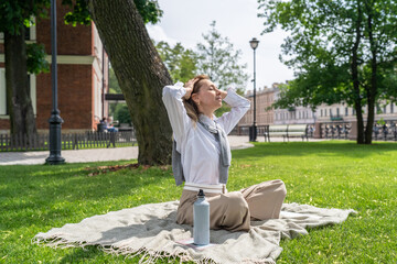 Relaxed smiling woman stretch arms facing sun, resting on grass in hot summer with water bottle in park. Dehydration prevention, sun protection, warm weather fatigue relief, skin care under UV