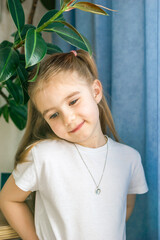 Smiling Little Girl in White Clothes with a Necklace Posing Near Houseplant, Looking at the Camera.
