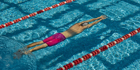 A competitive swimmer performs a backstroke in a sunlit pool lane, with clear blue water sparkling...