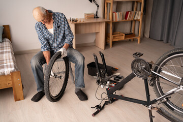 Retired man in casual clothes adjusting bicycle wheel at home in the living room interior. Bike...