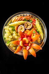 Assorted fresh fruits including grapes, strawberries, pears, orange peel, apricots, and passion fruit, beautifully arranged on a silver plate against a black background.