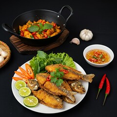  plate of cá chiên giòn, crispy fried fish with fish sauce dip, on a black background, garnished with fresh herbs and lime wedges. A hot wok and a coastal vendor’s tray