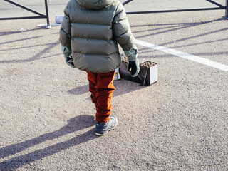 Child in winter clothing stands in a parking lot observing a used fireworks box in daylight, possibly after a New Year celebration event.