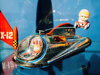 Colorful vintage tin space toy rocket labeled X-12 with astronauts in suits displayed in a museum setting against a vibrant blue background