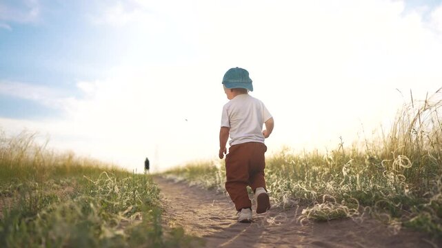 Child walking on dirt path. Toddler is walking on sunny path in field. A male exploring nature in a casual hat. A child strolls along a dirt trail lifestyle.