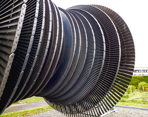 Close-up view of a large metallic turbine rotor with numerous evenly spaced blades, displayed outdoors on a gravel base with greenery and a building in the background