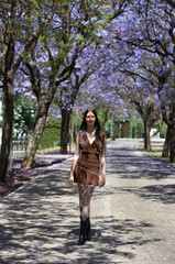 A young, attractive Spanish woman with dark hair, tattoos, long hair, a brown dress, and black boots walks on a spring day among large jacaranda trees in bloom.