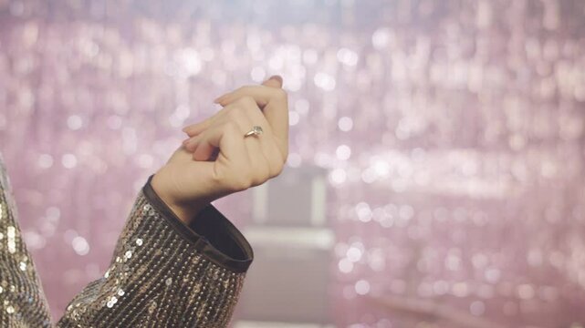 A close-up of a woman's hand, snapping her fingers, wearing a diamond ring and a silver sequin jacket, moving gracefully. The shimmering pink bokeh background creates a glamorous party atmosphere.
