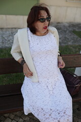 Elegant mature woman wearing white lace dress and sunglasses posing on bench