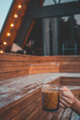 Hand holding a glass mug of iced coffee on a wooden terrace with cozy lights and a modern cabin in the background