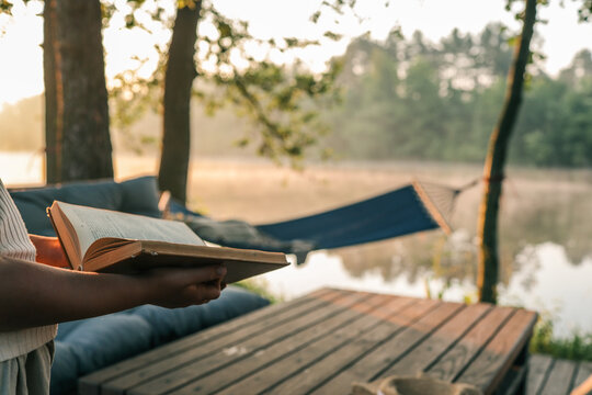 Person holding open book near lake at sunrise. Wooden terrace, hammock and trees create peaceful outdoor reading atmosphere in soft morning light