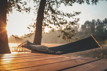 Morning sunlight casting warm rays on a wooden terrace with a blurred view of a hammock hanging between trees near a tranquil forest lake