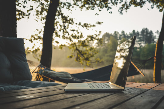 Laptop placed on a wooden table in a tranquil forest setting with a hammock and soft morning light, ideal for remote work or digital nomad lifestyle - Powered by Adobe