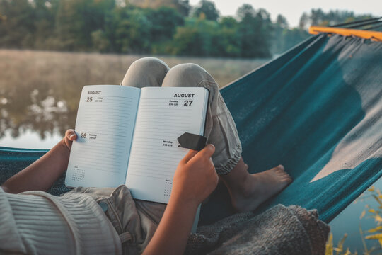 Person relaxing in a hammock by the lake, holding an open planner showing August dates, enjoying a peaceful moment outdoors