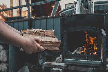 Person holding chopped firewood near an outdoor black metal stove with a burning fire inside,...