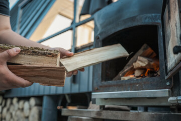 Person holding chopped firewood near an outdoor black metal stove with a burning fire inside,...