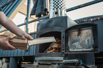 Person holding chopped firewood near an outdoor black metal stove with a burning fire inside,...