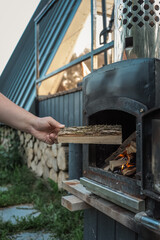 Person holding chopped firewood near an outdoor black metal stove with a burning fire inside, located on a wooden terrace near a modern cabin