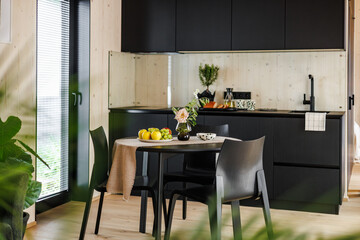 Bright Scandinavian-style kitchen with black cabinetry, a cozy dining table featuring fresh fruit, floral centerpiece, and dotted ceramics in a minimal modern interior