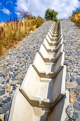 Concrete drainage channel on a hillside lined with stones surrounded by dry grass and shrubs under a blue sky