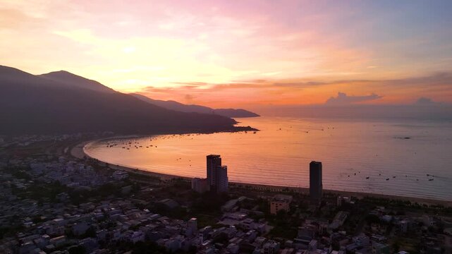 Golden sunlight bathing da nang coastline, fishing vessels anchored near shoreline, modern skyline contrasting verdant mountain landscape during serene twilight moment