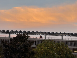 Silhouetted pedestrians with bicycle traverse an elevated modern walkway beneath a dramatic orange...