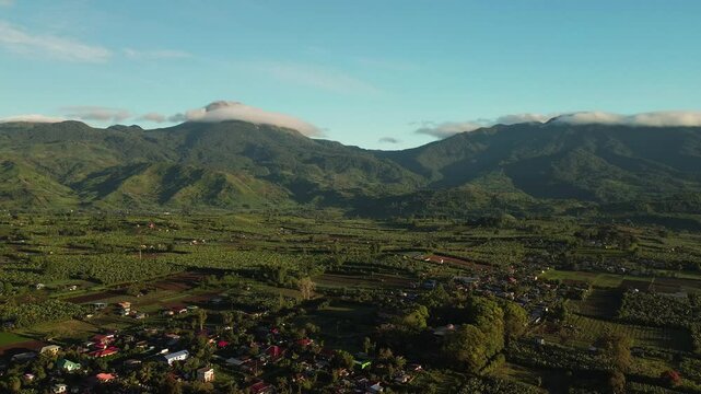 Wide aerial panorama reveals peaceful villages, patchwork fields, and abundant greenery at the foot of cloud-capped Mount Apo, bathed in gentle morning sunlight in Davao, Mindanao.