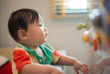 Baby stands up while holding onto playpen wall and looking at the toy stick on the cabinet with natural light