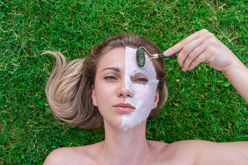 Woman's face with cosmetic face mask and with roller smoothing wrinkles in grass. Spa, healthy,...