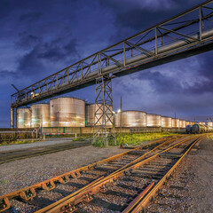 Industrial scene whit rails on the foreground, shiny silos on the background and al large pipeline...