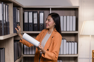 Businesswoman is searching files in the archive of her office, holding some documents and smiling,...