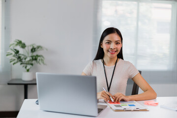 Young Asian businesswoman analyzing financial data on a laptop while taking notes on a clipboard, working diligently at her modern office desk with natural light streaming in