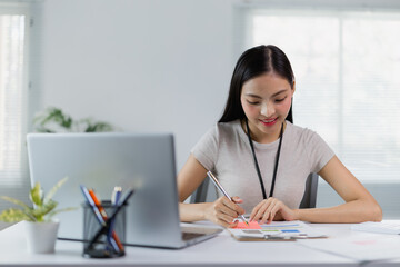 Young Asian businesswoman smiling and working with financial documents using laptop and taking notes on a sticky note while sitting at her desk in a modern office