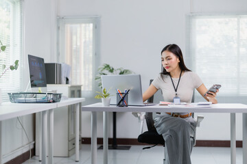 Young Asian businesswoman using laptop and holding smartphone while sitting at her desk in modern...