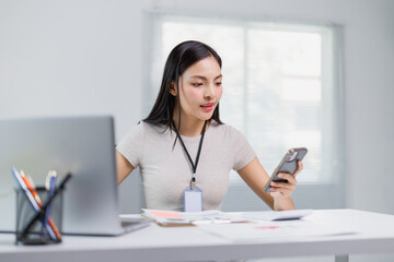 Young office employee wearing an ID card, using a smartphone while working on a laptop and managing documents on a desk in a modern, well-lit office environment